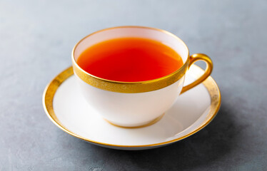Elegant cup of amber tea in a fine china cup with gold trim, set on a saucer. Grey background. Close up.