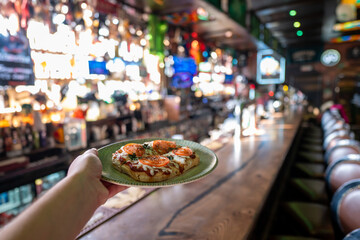 A person's hand holds a small plate of tasty pizzetta or bruschetta appetizers in a dimly lit bar or pub interior with a long wooden counter.