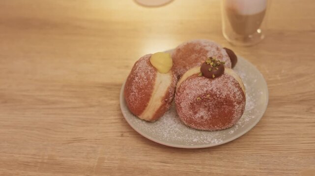 Slow camera movement showing a woman picking a sugar-coated filled doughnut from a plate, with a cappuccino in the background