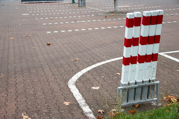 Red and white striped traffic bollards stand on a paved parking lot, surrounded by fallen leaves, marking the edge of a designated area for vehicles and pedestrians