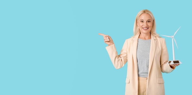 Mature businesswoman with wind turbine model pointing at something on blue background with space for text