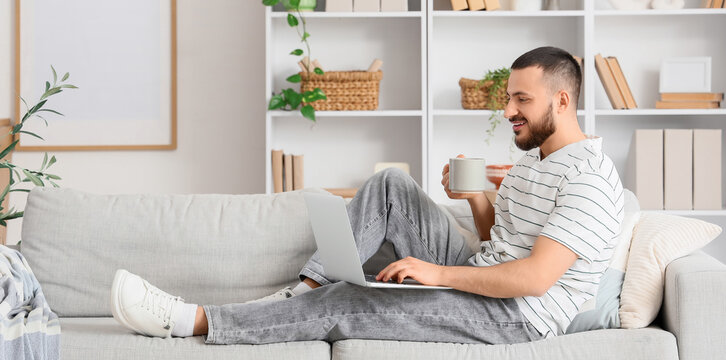 Young man with coffee using laptop while sitting on sofa at home
