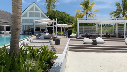 Luxury resort photo of Maldives tropical paradise island in Indian Ocean with swimming pool, palm trees, beach umbrella, chair. Beautiful photography, wooden house, sky with clouds.
