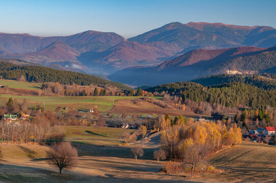 View of undulating fields and colorful forests blanket the landscape beneath majestic mountains fading into a soft blue sky, Povraznik, Banska Bystrica Region, Slovakia.