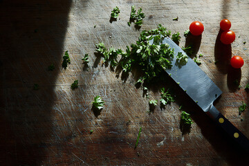 Close up overhead view of a chef s knife chopping fresh green herbs and cherry tomatoes on a rustic wooden cutting board with dramatic lighting