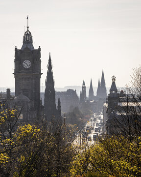 View of the Balmoral clock tower and gothic spires punctuate the skyline above a bustling street framed by spring foliage, Edinburgh, Scotland, United Kingdom.