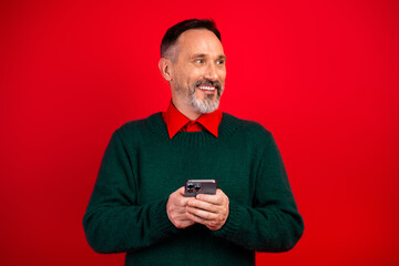 Funny merry man in green sweater smiling at his phone against a red xmas background and festive vibe that feels warm and humorous