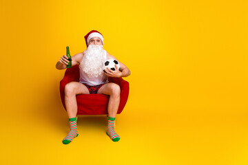 Cheerful Santa relaxes at home in a red chair with beer and soccer ball under a bright yellow background