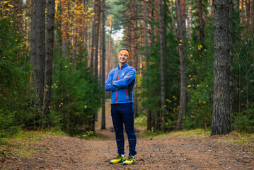 Naklejka premium Man standing on a dirt trail in a forest, smiling, dressed in sportswear with arms crossed, enjoying an active outdoor lifestyle and healthy fitness routine during autumn season