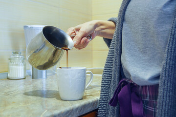 Woman in a home outfit pouring freshly brewed Turkish coffee from a cezve into a white ceramic mug. Represents morning ritual completion, handcrafted beverage serving, and mindful breakfast moment.