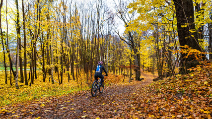 Middle-aged woman riding bicycle on path in forest in autumn. Back view