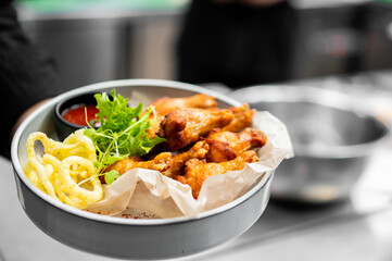 A close-up, horizontal shot of golden, crispy fried chicken wings, served in a gray bowl lined with parchment paper, accompanied by curly fries, fresh lettuce garnish, and a red dipping sauce
