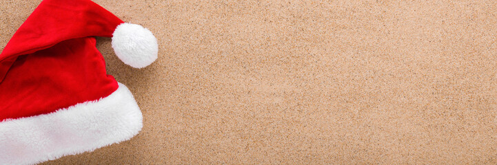 Bright red hat with white fur on light brown dry sea sand background. Wide banner. Closeup. Garment for Christmas celebration. Empty place for text. Top down view. © fotoduets