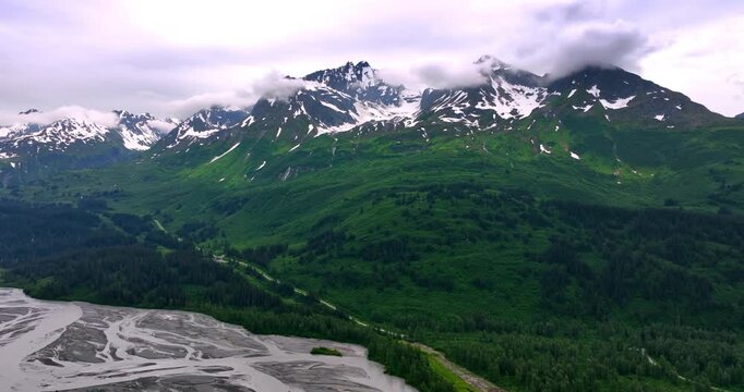 Clouds hide the peaks of the mountains covered with snow at tops and greenery at the foot. Branching river flows in the valley. Alaska, USA. Aerial view.
