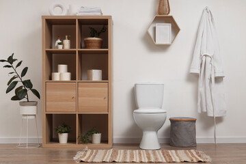 Interior of light restroom with toilet bowl, shelf unit and bathrobe