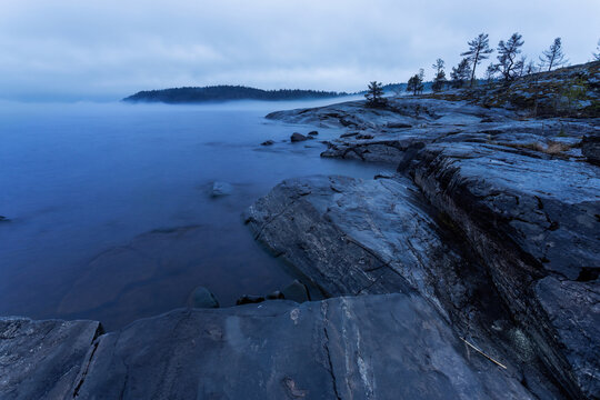 View of dark, striated rocks meeting the misty, ethereal water under a cloudy sky, a shoreline fading into the distance, Sortavala, Russia.