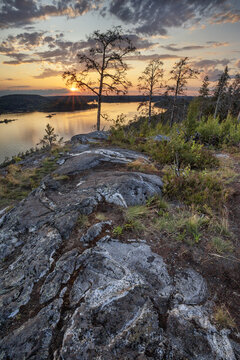 View of the sun dipping below the horizon, as seen from a rocky outcrop overlooking a calm lake, with silhouetted trees against the colorful sky, Sortavala, Russia.