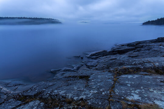 View of fog rolling over the still, dark waters, meeting the rocky shoreline under a cloudy sky, shrouded in mystery, Sortavala, Russia.
