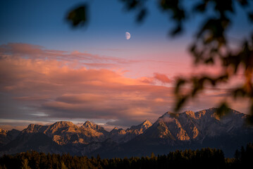 View of the majestic mountains under a sky brushed with fiery hues as twilight embraces a crescent moon, framed by soft foliage, Nesselwang, Bayern, Germany.