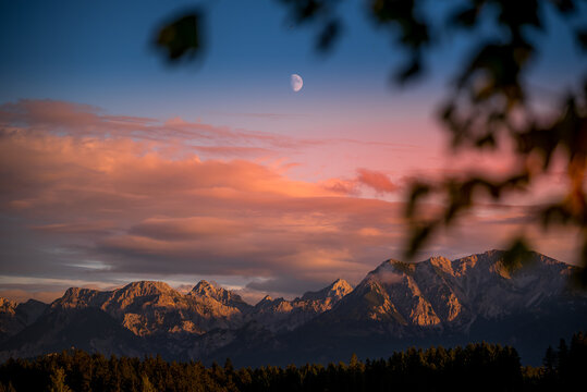 View of the majestic mountains under a sky brushed with fiery hues as twilight embraces a crescent moon, framed by soft foliage, Nesselwang, Bayern, Germany.