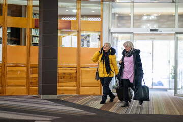 Two older women dressed in winter clothing standing in a hotel lobby and having a friendly conversation next to their luggage. Concept of travel, friendship, and leisure lifestyle for seniors.