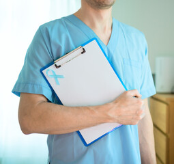 Close up of a male doctor in medical uniform holding clipboard with blue ribbon and empty sheet. November prostate cancer awareness month symbol of prevention and early diagnosis support.