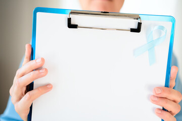 Close up of a young medical worker holding clipboard with blank paper and blue ribbon. November symbol of prostate cancer awareness, prevention, and healthcare support.