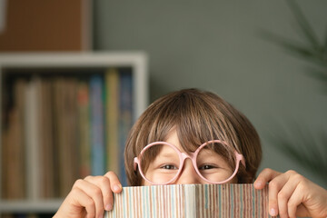 A little boy wearing round pink glasses peeks from behind a fascinating book at table. Joyful sly look at camera showing anticipation of reading and imagination growth.