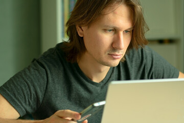 A young man with long hair sits at his desk working intently on a laptop. His gaze is focused on the computer monitor. Home office and remote employment concept.