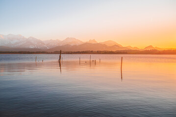 View of a serene lake mirroring the warm hues of the setting sun, with snow-capped mountains fading into the horizon, Hopfen am See, Fussen, Bavaria, Germany.