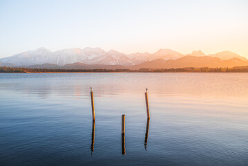 View of serene lake waters reflecting distant, snow-capped mountains under a soft, warm golden light, Hopfen am See, Bavaria, Germany.