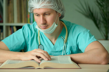 A male doctor or nurse in medical uniform with mask and cap sits at his desk reading a book. Stethoscope around his neck. Studying scientific literature and medical education concept.