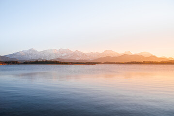 View of a tranquil lake mirroring distant, snow-capped mountains under a soft, golden sunset glow, casting a serene ambiance, Fussen, Bavaria, Germany.