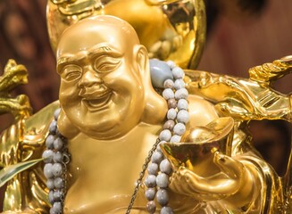 Close up of a golden Laughing Buddha statue (Budai or Hotei) holding a gold ingot. Figure symbolizes wealth, happiness, and good fortune in Feng Shui and Buddhism.