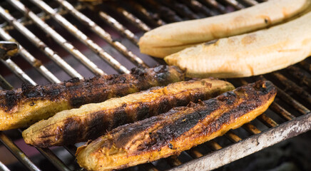 Close up of ripe plantains roasting on a hot metal barbecue grill. The skin is charred and...