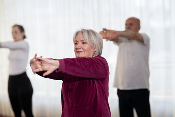 Active elderly woman in sportswear stretching her arms forward during a group fitness or rehabilitation session. Concept of healthy aging, wellness, flexibility, and active senior lifestyle.