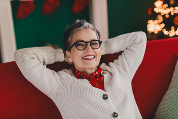 Fototapeta premium Smiling senior woman in cozy winter cardigan relaxing at home near festive Christmas decorations and a lit-up tree