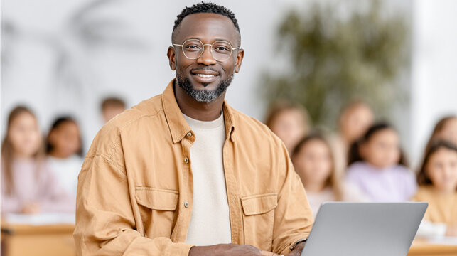 African American man in a casual tan jacket is smiling while working on a laptop in a classroom filled with engaged students, showcasing a collaborative learning environment