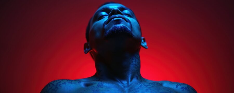 Man tilts head back eyes closed bathed in dramatic blue and red neon light. Chest tattoos visible under vibrant illumination. Studio portrait conveys mood of peace or contemplation.