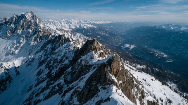 Breathtaking aerial view of majestic snow-covered mountain peaks and sharp ridges with brown rocky slopes, deep valleys, and distant mountain ranges under bright blue sky.