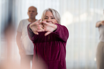 Fototapeta premium Active elderly woman in sportswear stretching her arms forward during a group fitness or rehabilitation session. Concept of healthy aging, wellness, flexibility, and active senior lifestyle.
