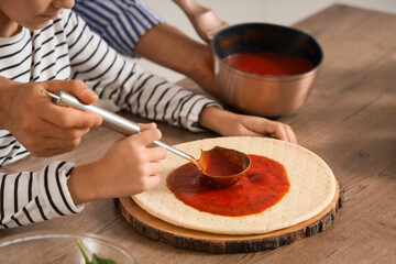 Father with his daughter applying tomato sauce to pizza dough at table in kitchen, closeup