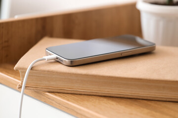 Mobile phone with charging cable and book on shelf in room, closeup