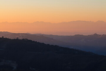 Stunning evening sunset over Piedmont's Langhe and Asti region, with the majestic snow-capped Monte Rosa and Alps glowing in warm alpenglow against a dramatic pink-orange sky. Perfect panoramic vista 