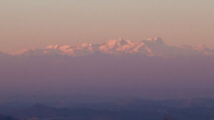 Stunning evening sunset over Piedmont's Langhe and Asti region, with the majestic snow-capped Monte Rosa and Alps glowing in warm alpenglow against a dramatic pink-orange sky. Perfect panoramic vista 