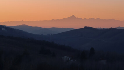 Stunning evening sunset over Piedmont's Langhe and Asti region, with the majestic snow-capped Monte Rosa and Alps glowing in warm alpenglow against a dramatic pink-orange sky. Perfect panoramic vista 