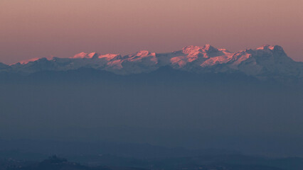 Stunning evening sunset over Piedmont's Langhe and Asti region, with the majestic snow-capped Monte Rosa and Alps glowing in warm alpenglow against a dramatic pink-orange sky. Perfect panoramic vista 