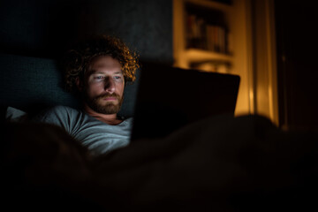 A man with curly hair is sitting on a bed and looking at a laptop