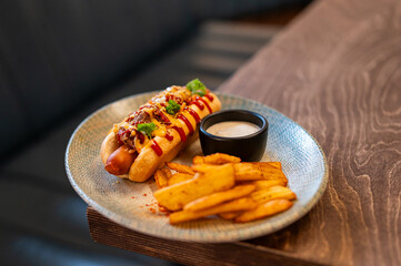 A gourmet hot dog topped with chili, ketchup, mustard, and herbs, served with potato wedges and a dipping sauce on a plate over a wooden background