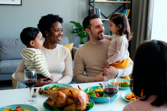 Multiethnic family sitting at dining table, Black woman holding toddler boy sharing meal together, visible roast turkey and wine glasses